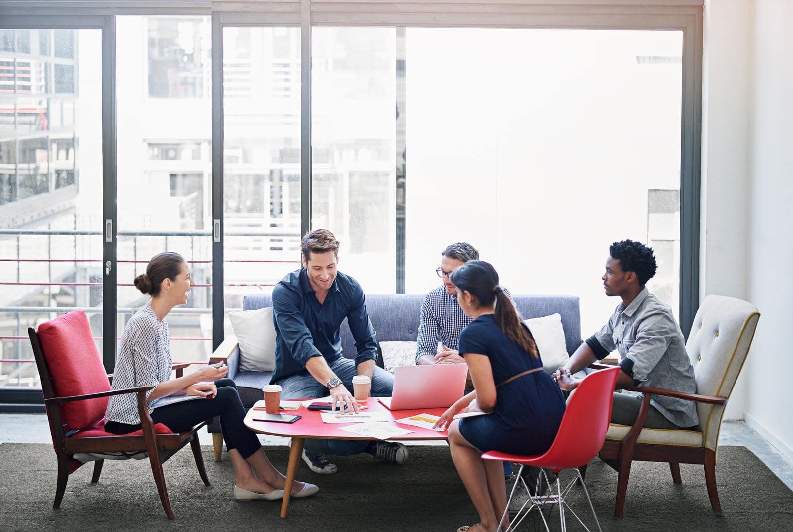 A group of five people is sitting around a small red coffee table in a modern office setting. They appear engaged in a discussion, with documents, a laptop, and coffee cups on the table. The room features large windows, letting in natural light, and the seating includes a mix of chairs and a sofa, creating a casual meeting atmosphere.