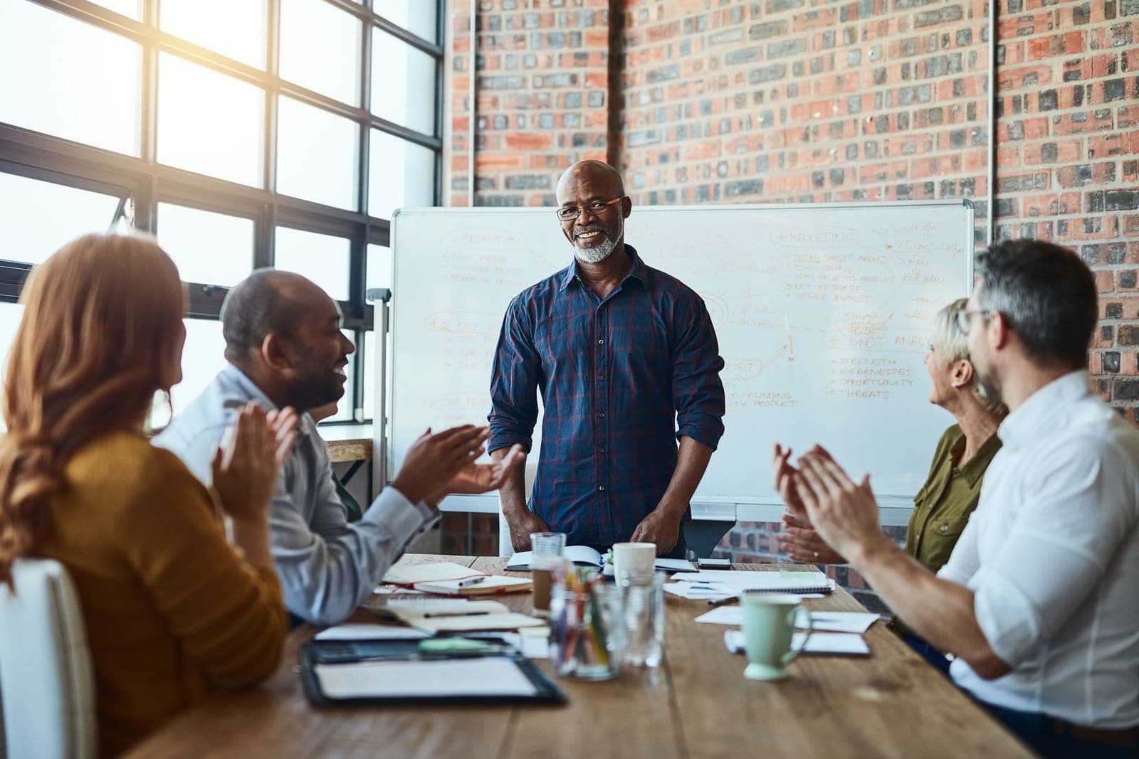 49699026_what-a-great-way-to-start-a-project-a-group-of-businesspeople-in-the-office-applauding-a-colleague-after-a-successful-group-meeting A group of five people is gathered in a modern office setting with a brick wall and large windows. A man in a plaid shirt stands in front of a whiteboard, smiling, while others sit around a wooden table with notebooks, mugs, and documents. Two people are clapping, and the overall atmosphere is positive and collaborative.