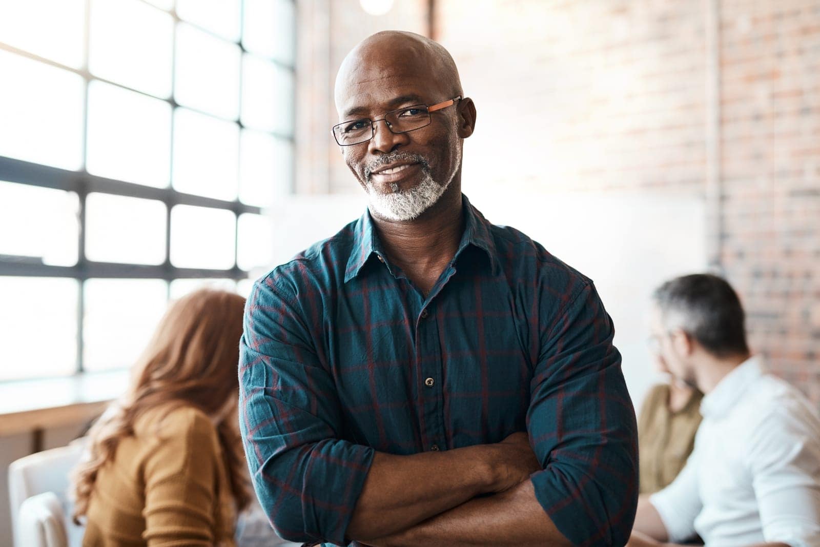 A man with glasses and a graying beard stands confidently with crossed arms, wearing a checked shirt. He is in a bright loft-style office with large windows and exposed brick walls. In the background, a woman with long hair and a man with glasses are seated and engaged in conversation.