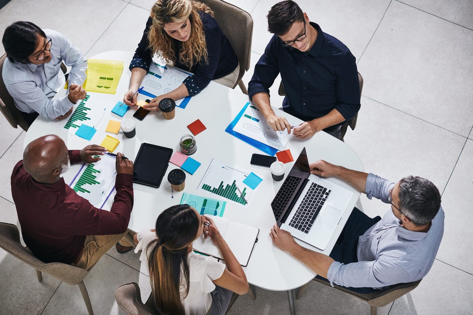 53430076_meeting-discussion-and-top-view-of-a-team-planning-in-a-company-report-or-proposal-in-the-office-teamwork-diversity-and-marketing-employees-working-on-a-creative-project-together-in-the-workplace A group of six people are gathered around a white table with a laptop, documents, and colorful sticky notes. They appear engaged in a discussion or meeting, with graphs and charts visible on paper. There are coffee cups and a small plant on the table. The setting is professional, likely an office or conference room.