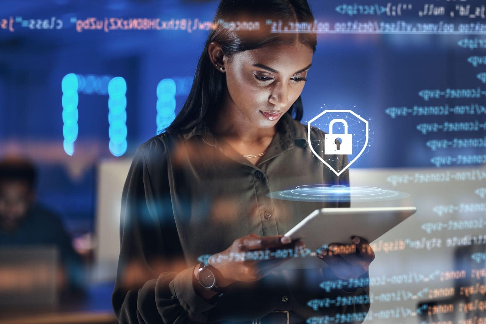 A woman is intently using a tablet, with a digital overlay of code and a security lock icon in the foreground. The atmosphere is that of a tech-oriented environment with blue lighting and another person blurred in the background at a computer. She wears a dark blouse and a watch, focusing on the device in a high-tech setting.