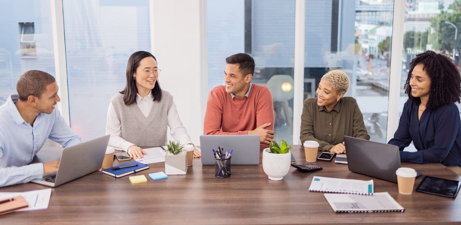 A group of five people are sitting around a conference table in a modern office setting. They are engaged in a friendly meeting, with laptops, notebooks, and coffee cups in front of them. The room has large windows with a view of an urban landscape in the background. The atmosphere appears professional and collaborative.