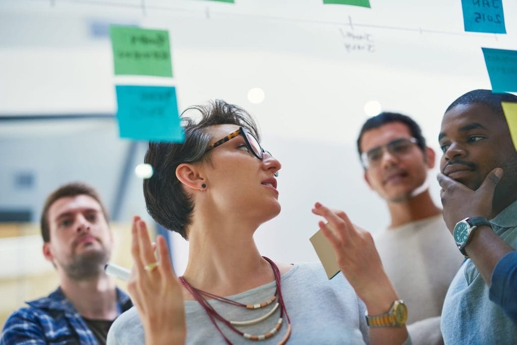 A group of four colleagues stands in a modern, bright office, gathered around a glass wall covered with colorful sticky notes and a sketched timeline. A woman with short hair and glasses speaks and gestures while holding a pen and a note, as three men listen—one resting his hand on his chin thoughtfully. The scene suggests a collaborative brainstorming or planning session.