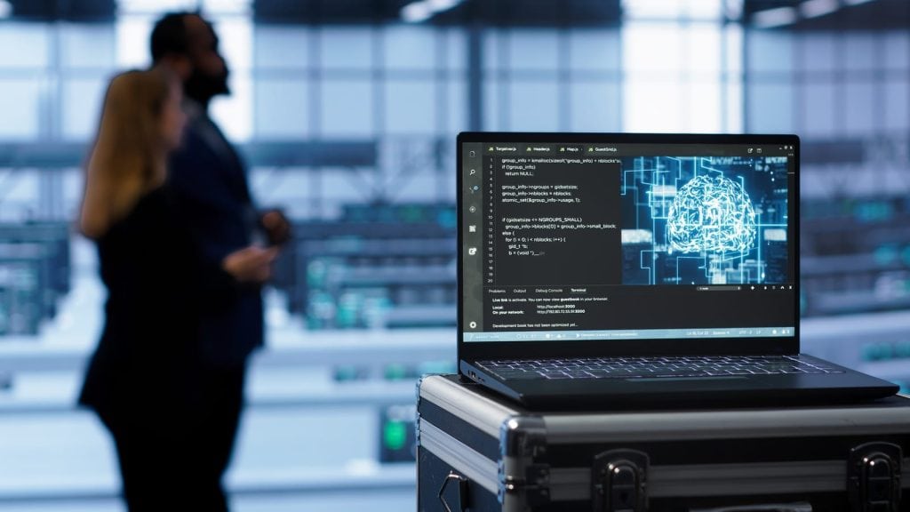 A laptop sits on a metal equipment case in a modern, glass-walled tech facility or data center. Its screen shows a dark-themed code editor with programming text alongside a glowing, neon-blue digital brain graphic, suggesting AI or machine learning. In the blurred background, two business-dressed people converse amid rows of workstations or machinery with small green indicator lights. The scene is bright and industrial, with large windows and a high ceiling. No recognizable brands or landmarks are visible.