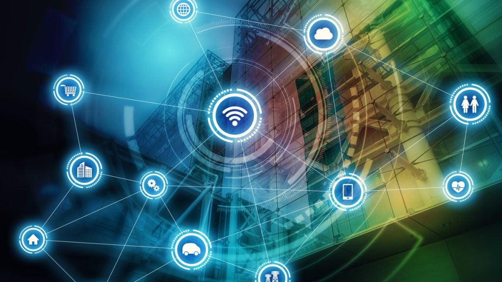 A futuristic, tech-themed illustration showing an Internet of Things network overlaying a modern glass building. In the center is a glowing Wi‑Fi symbol, with lines connecting to circular icons around it: a cloud, smartphone, shopping cart, people, heart/health, car, home, factory/city buildings, and gears. The color palette is blue and green, with HUD-style graphics and interconnected nodes suggesting digital connectivity across services and devices.