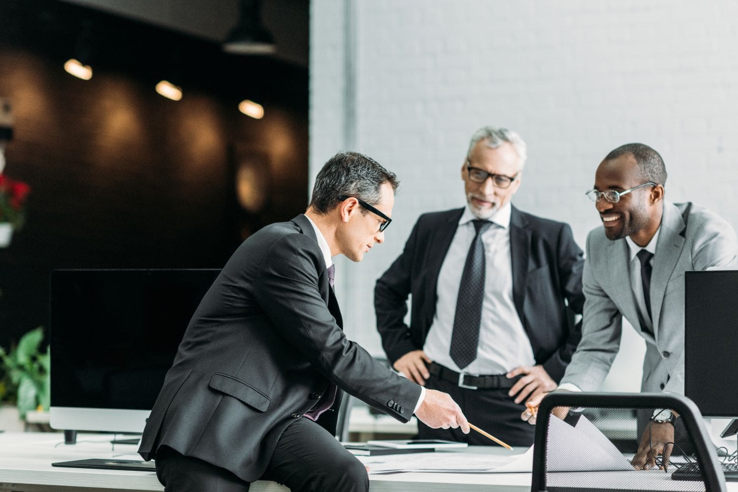 Three businessmen in suits collaborate in a modern office. One, seated on the edge of a desk, points at documents with a pencil while two colleagues stand nearby, listening and smiling. Large computer monitors sit on the desks, with a white brick wall and a darker, softly lit background with plants. The scene conveys a professional meeting and discussion.