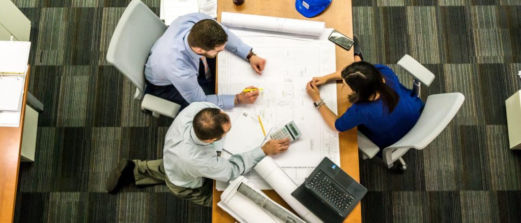 Top-down view of three colleagues collaborating at a conference table in an office. They are studying large architectural/engineering blueprints with pencils while one uses a calculator. Rolled plans, a laptop, a smartphone, and a blue hard hat sit on the table. The group is seated in gray office chairs on a striped carpet, with binders and desks at the edges of the scene.