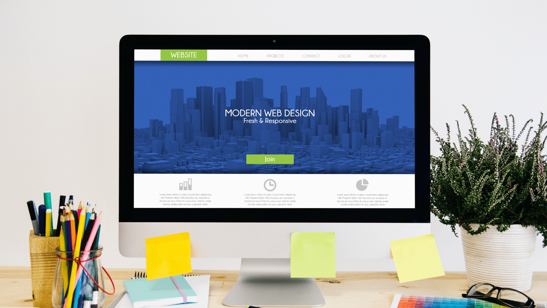 A clean, creative workspace with a large desktop monitor on a wooden desk. The screen shows a modern website landing page featuring a blue 3D cityscape, the title “MODERN WEB DESIGN – Fresh & Responsive,” a green “Join” button, and a top navigation bar with Website, Home, Projects, Contact, Log In, and About Us. Sticky notes are on the monitor stand. On the left are jars with colored pencils and pens, plus notebooks and more sticky notes. On the right sits a leafy plant in a white pot and a pair of eyeglasses beside a color chart. The background is a plain light wall.