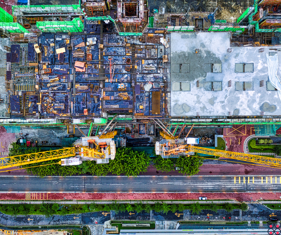 A top-down aerial view of a large urban construction site. The frame shows a partially built multi-story structure with exposed rebar, concrete slabs, formwork, scaffolding, and green safety netting. Two yellow tower cranes extend from the middle toward both sides. Below the site runs a divided roadway with lane markings, a crosswalk, a few vehicles, and landscaped medians lined with trees. The scene is dense with building materials, equipment, and geometric patterns created by the grids of the construction and surrounding streets.