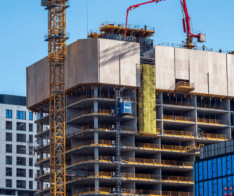 A high‑rise building under construction with multiple exposed concrete floors and yellow safety railings. A tall yellow tower crane stands beside the structure, while a red boom concrete pump works at the top among temporary formwork and panels. A vertical construction hoist with a blue car runs up the façade next to a section wrapped in green mesh. Surrounding the site are other modern buildings, including a blue‑glass structure on the right, under a clear blue sky.
