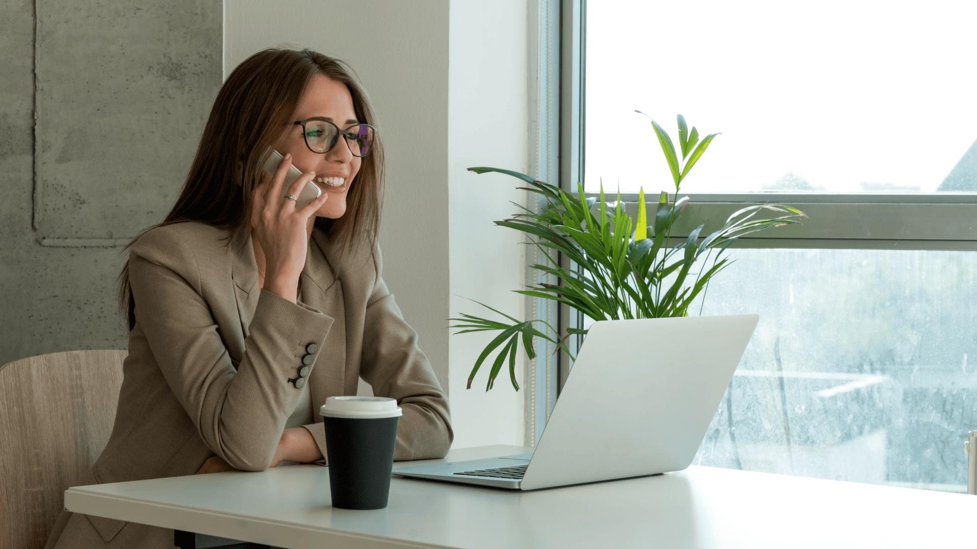 A smiling woman in a beige blazer and glasses sits at a modern office desk, talking on a smartphone. An open silver laptop is in front of her beside a black takeaway coffee cup. A green potted plant sits near a large window that lets in daylight, with a concrete wall and a wooden chair completing the minimal workspace. No recognizable brands or landmarks are visible.