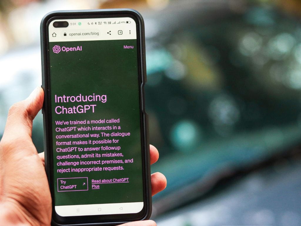 A person holds a smartphone outdoors, displaying the OpenAI website on the screen. The page shows the OpenAI logo and the headline “Introducing ChatGPT” in purple text on a dark background, with buttons labeled “Try ChatGPT” and “Read about ChatGPT Plus.” The URL bar shows openai.com/blog, and the background is blurred with bokeh lights.