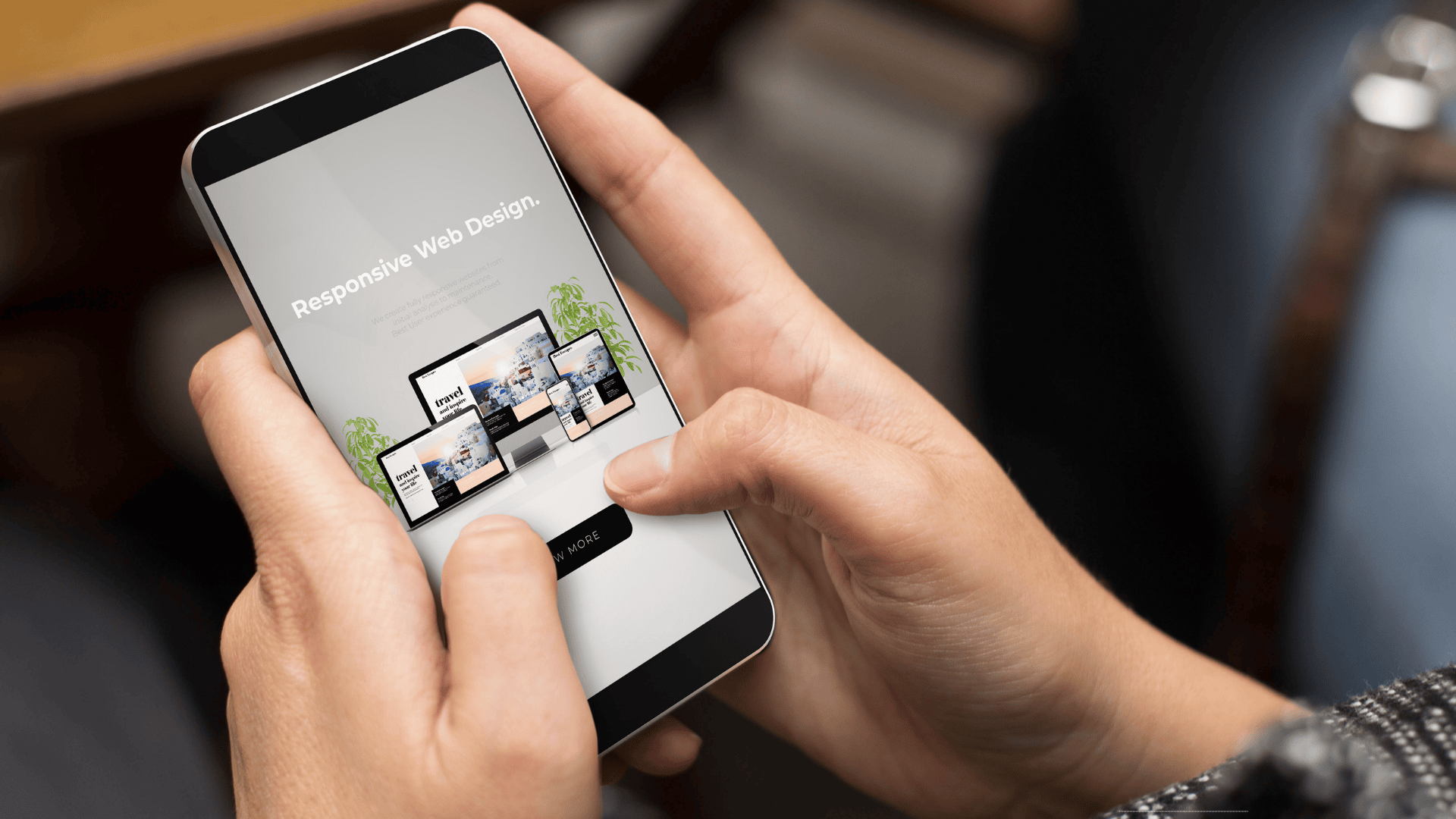 Close-up of a person holding a smartphone, viewing a landing page for “Responsive Web Design.” The screen shows device mockups (desktop, tablet, and phone) displaying a “travel magazine” site, with a black “VIEW MORE” button below. The background is softly blurred, suggesting an indoor setting; a wristwatch is visible out of focus in the upper right.
