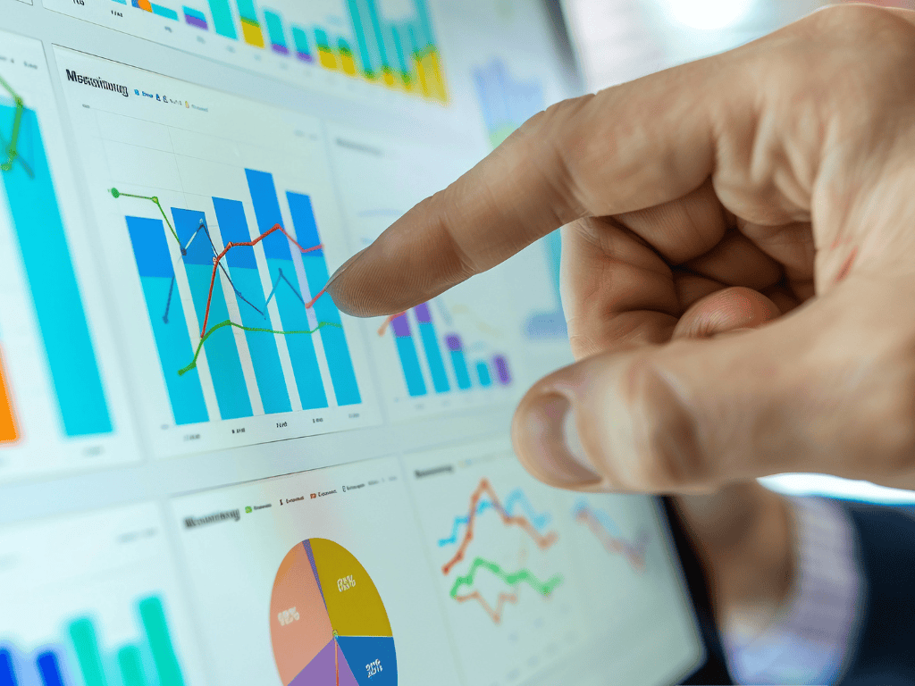 Close-up of a person’s hand in a suit pointing at a touchscreen displaying a business analytics dashboard with multiple panels of colorful bar charts, line graphs, and a pie chart showing trends and metrics.