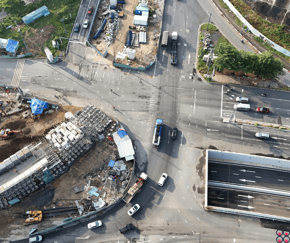 Aerial view of a large multi-lane intersection with active road construction. Cars, vans, motorcycles, and several trucks move through the junction. Two corners are construction zones with scaffolding, stacks of concrete blocks, blue tarps, containers, pipes, and an excavator. At the lower right is a newly built overpass or bridge deck with fresh asphalt and directional arrows. A fenced site and greenery line the upper-right side. No recognizable figures, landmarks, or brands are visible.