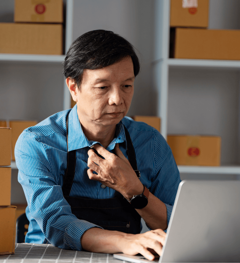 A middle‑aged man in a blue striped shirt and dark apron sits at a desk using a silver laptop, one hand adjusting his collar. He wears a black smartwatch. Around him are shelves and stacks of brown shipping boxes with red stickers, and a grid cutting mat on the table, suggesting a small business packing or home office workspace.
