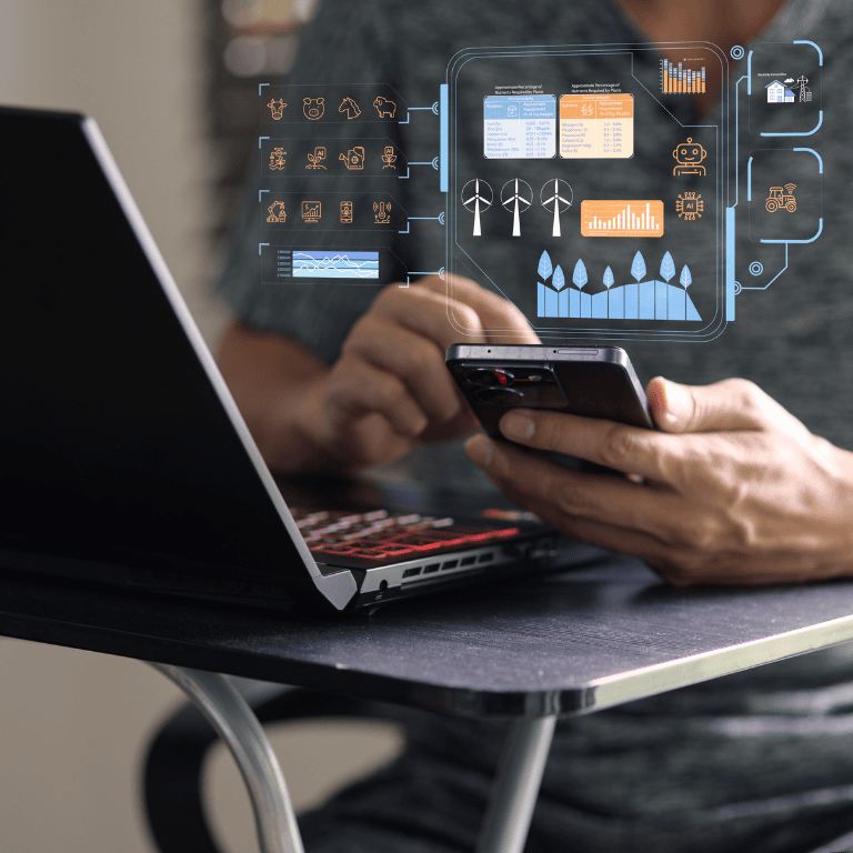 A person sits at a small desk using a smartphone in one hand and a laptop with a red‑backlit keyboard in front of them. Overlaid holographic graphics show a digital dashboard with charts and data visualizations, including wind turbines, bar graphs, line graphs, plant and leaf icons, a robot/AI symbol, a house, a tractor, and various IoT and weather icons. The scene suggests remote monitoring or smart analytics for energy and agriculture in a modern home or office setting.