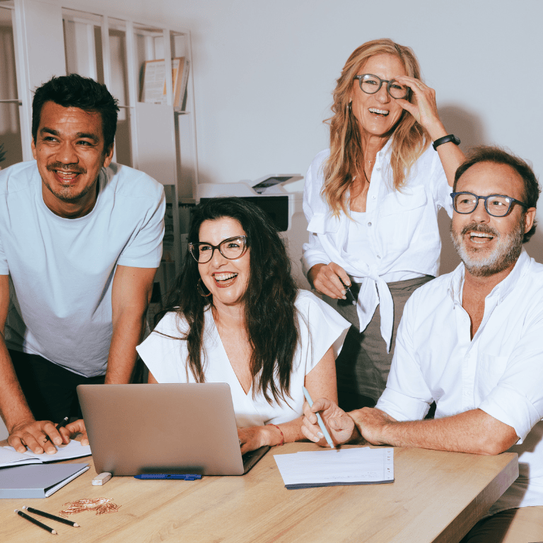 Four colleagues in a bright office gather around a wooden table, smiling and laughing. Two women and two men, most wearing glasses, lean in toward an open laptop with notebooks, papers, pens, and a phone on the table. Shelving and a printer are in the background against white walls, suggesting a casual, collaborative meeting.