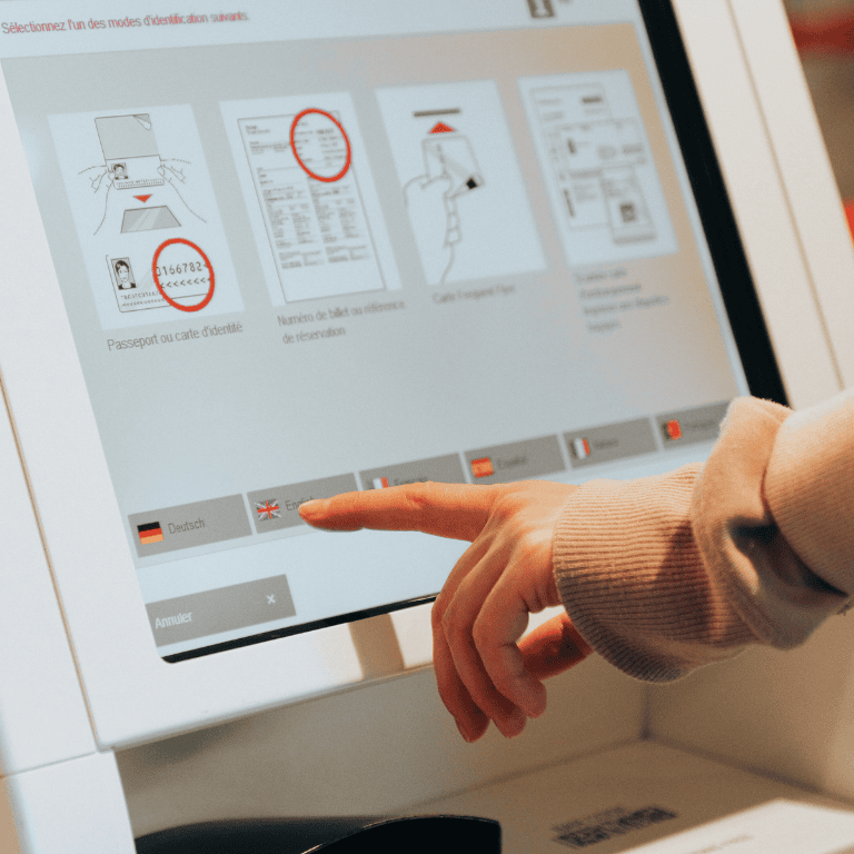 A close-up of a hand using a touchscreen self-service kiosk, tapping the “English” language button with a UK flag. The interface text is in French and displays options for identification: passport or ID card, ticket number or reservation reference, and frequent flyer card. Other language buttons (German, Spanish, French) and a “Annuler” (Cancel) button are visible along the bottom of the screen.